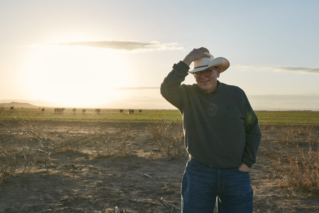 Roy Jackson stands in an open field at sunset, tipping his cowboy hat with a warm smile, representing his welcoming approach to business consulting and management.